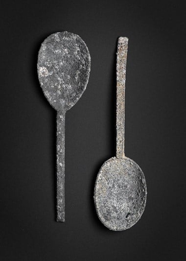 Two pewter spoons. The one on the left has a round maker’s mark and the initials “BA” at the base.