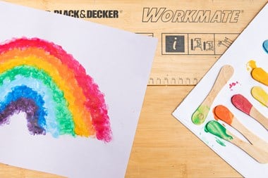 On the left hand side, a splotchy rainbow on a piece of white paper sits on top of a Black and Decker workboard. On the right hand side, a row of lolly sticks sit on an olod white envelope, with one end dipped in paint, each one representing a colour in the rainbow.