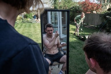 A topless young man, sat in a back garden, inspects his newly cut hairstyle in a long mirror. In front of him, a middle aged man mows the lawn and a woman beside him holds a pair of scissors.