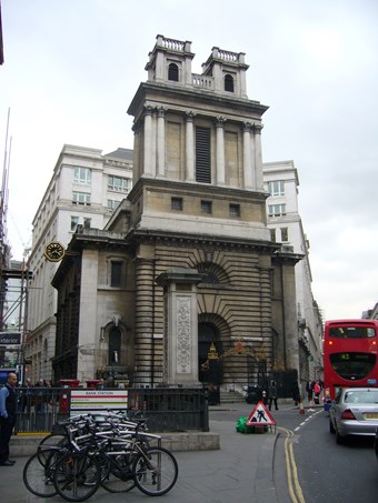 Imposing Baroque church of unusual design, on the corner of a busy London street. A red bus to right. The entrance bay is framed by plain columns, with deep rustication. Above, the broad rectangular tower has tall columns, and two square turrets.