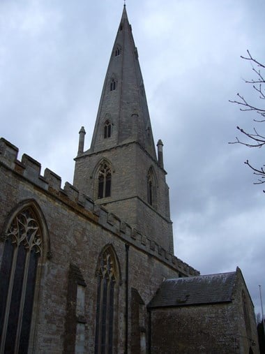 The west end of a church with a tall 14th-century pointed broach spire. The aisle wall in the foreground has pointed Gothic windows with tracery, and battlements. A stormy sky.