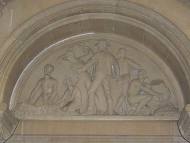 Stone tympanum, carved with figures in relief. Centre, a slave master stands holding a whip. Left, a woman and child kneel by a heap of cotton; behind a man carries a basket on his shoulders. Right, a man kneels emptying cotton from a basket.