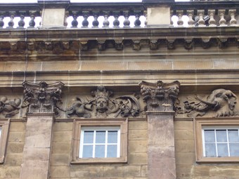 Carved frieze at the top of an 18th-century building, with Corinthian pilasters below a balustraded parapet. At centre, an African woman wearing a feather headdress, surrounded by swags of fruit, tusks, and a parasol. An elephant head to right.