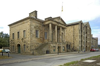 An 18th-century ashlar building in Classical style. It has a portico with four columns above a rusticated basement, with steps to either side. A flag flies from the pediment. Beside it to right is a tall rubblestone warehouse, of similar date.
