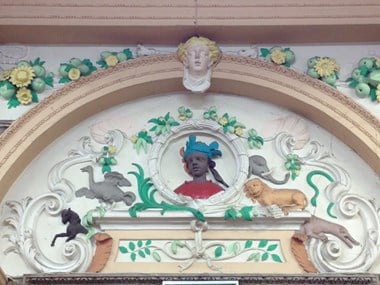 Tympanum with Rococo plasterwork, colourfully painted. Central roundel with head of a young African person in headdress. To left, an ostrich and a horse. To right, an elephant, lion and crocodile. Around, scrolled motifs with fruit, leaves and shells.