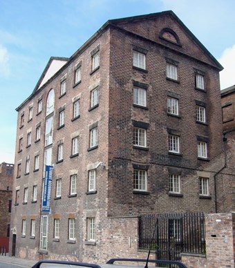 Tall 18th-century warehouse, of functional but elegant design, the red brick blackened in places. The street elevation has a pediment, below which the central bay has full-height windows below an arched window, where taking-in doors used to be.