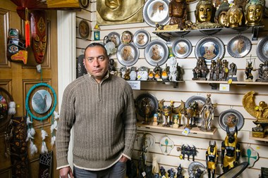 Portrait of a man, standing in shop in front of a display of ancient world artefacts. 
