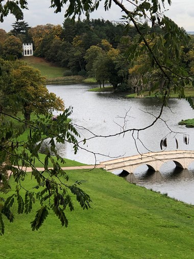 Round white building nestled in the woodland with a lake and arched bridge in the foreground