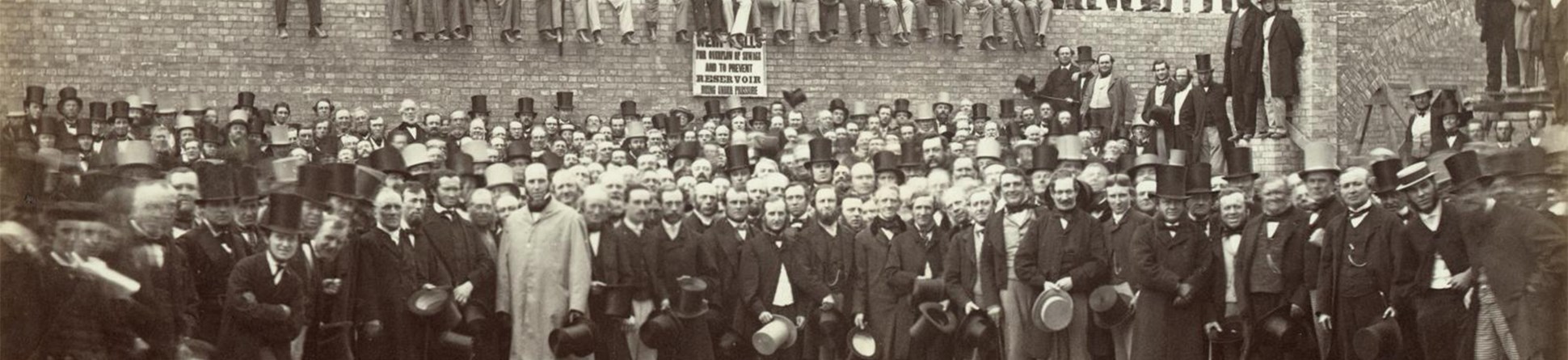 A large group of men posed in front of and on top of the brick wall forming part of the overflow reservoir at Crossness Pumping Station.  Most of the men are smartly dressed complete with top hats. A sign on the wall reads “WEIR WALLS FOR OVERFLOW OF SEWAGE AND TO PREVENT RESERVOIR BEING UNDER PRESSURE”.