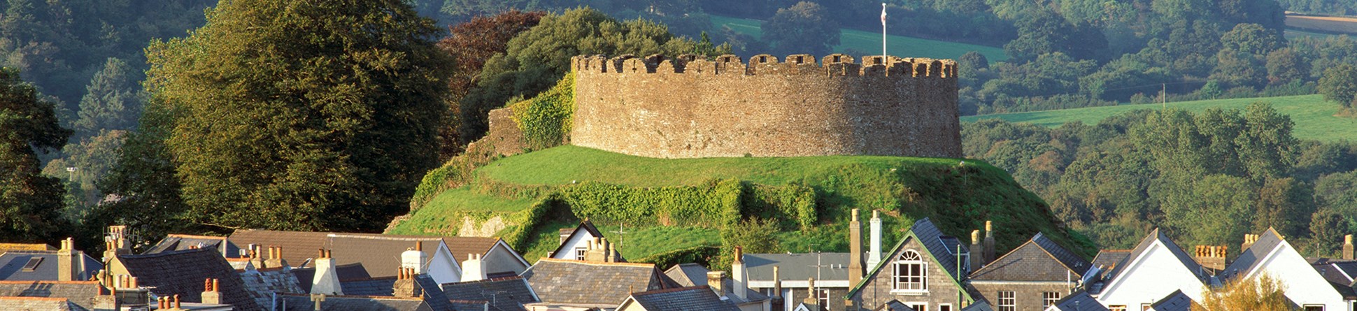 In the foreground is the pitched roofs of local houses, whilst in the background is Totnes Castle on a small hill, an example of a Norman motte and bailey castle. Rolling hills and woodlands can be seen in the background. Photo is taken in late spring or early summer.