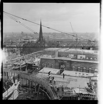 Black and white photograph of the Bull Ring construction site from a high vantage point. A crane extends across the image and builders can be seen at work. The old Market Hall is shown prior to its demolition.
