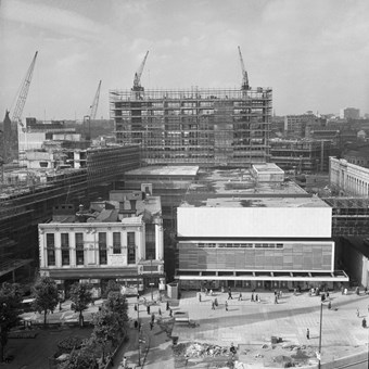 Black and white photograph showing the construction of the Bull Ring shopping centre from a high vantage point. Numerous buildings are being constructed, including a large office block. In the foreground, the new Woolworth store is shown next to the remaining part of the old store.
