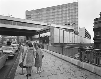 Black and white photograph taken from the street, showing part of the Bull Ring Shopping Centre. Two women in heavy coats are walking on the pavement alongside a busy road. A bridge spans across the road, beneath which is the district bank. An office block rises in the background.