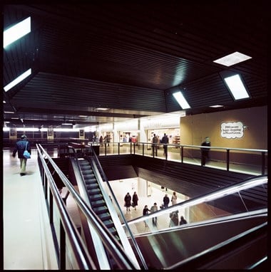 Colour photograph showing escalators and shoppers inside Wood Green Shopping City. A sign on the wall reads ‘1980 means happy shopping in Wood Green’.