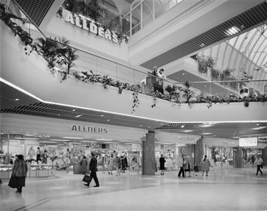 Black and white photograph showing the multiple levels of the Allders store at Eastgate Shopping Centre, including a balcony with plants. Shoppers are walking around leisurely.