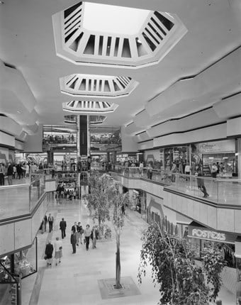 Black and white photograph looking along the mall at Queensgate Shopping Centre, showing the multiple levels with shoppers, plants, and roof lights.