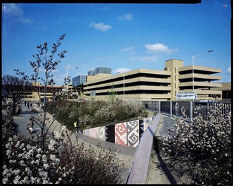 Colour photograph of the Queensgate shopping centre showing a concrete multi storey car park. In the foreground there is a pedestrian subway surrounded by plants in bloom.