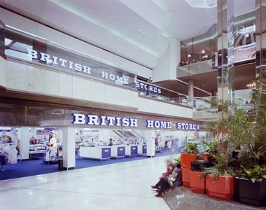 Colour photograph of the Queensgate Shopping Centre showing the interior entrance to British Home Stores. A large shop sign is above the entrance, and a till area can be seen inside the store. On the right, there is a seating area with green plants.