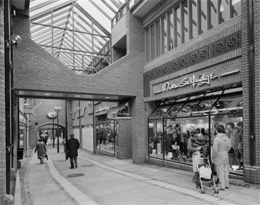 Black and white photograph showing one of the glass-covered lanes at The Lanes Shopping Centre, with shoppers walking through. On one side there is a Miss Selfridge store.