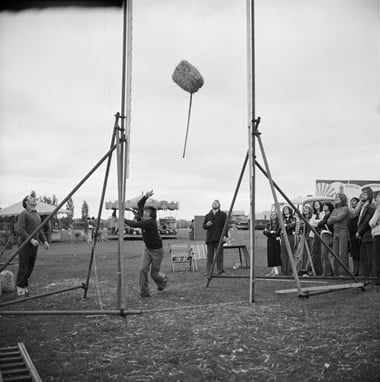 A man attempting to toss a hay bale over a pole with spectators watching on