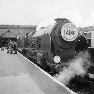 The engine of a steam train alongside a platform. On the front of the engine is a sign with the word ‘LAING’ beneath two crossed flags, and ‘STAFF OUTING CORONATION YEAR’ around the edge. Steam rises from beneath the engine.