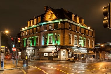 Large detached four-storey building lit up at night with white and green light. There are people sitting around tables outside on the right hand side of the building. There's a road with traffic lights, street lights and traffic signs on the foreground. In the background there is dark sky.