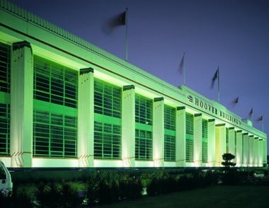The famous Art Deco Hoover Building lit up in green light at night time. There is a vegetation border in the foreground while the background shows a dark blue sky.
