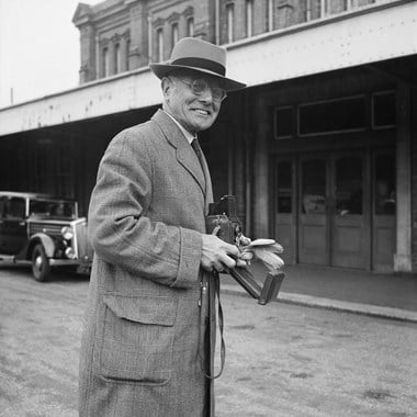 An old man smiling. He is holding a camera and wears an overcoat and hat.