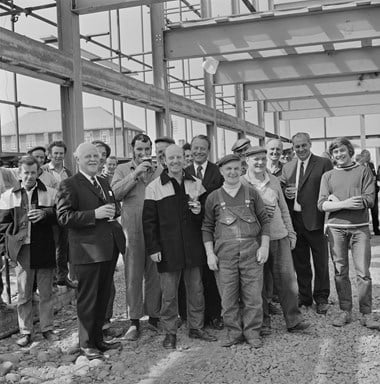 A crowd of men pose for a picture. Some are wearing work-overalls and donkey jackets and a few of the men wear suits. Some of the men are holding pints of beer. They look to be celebrating inside a large building which has scaffolding on the outside of it.