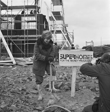 A woman plunges a shovel into the earth.  Next to her is a sign saying ‘Superhomes For the Money Programme.’ Behind her there is scaffolding around a half-built house. The image is in black and white.