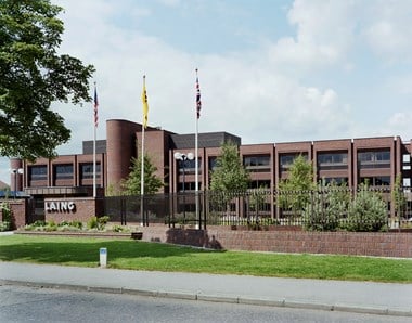 An exterior view of a large brick-coloured building. Three flags stand out the front. A Laing sign is posted on the front of the building. In the foreground, there is a hedge and a tree.