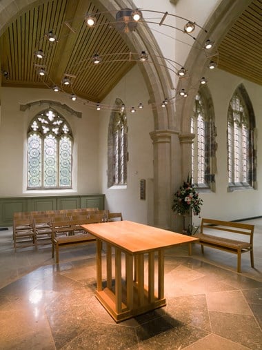 Interior of a church. The wooden furniture include a table in the centre with seats on the left and right area. The ceiling features a suspended circular light fitting. There are four patterned windows in the background.