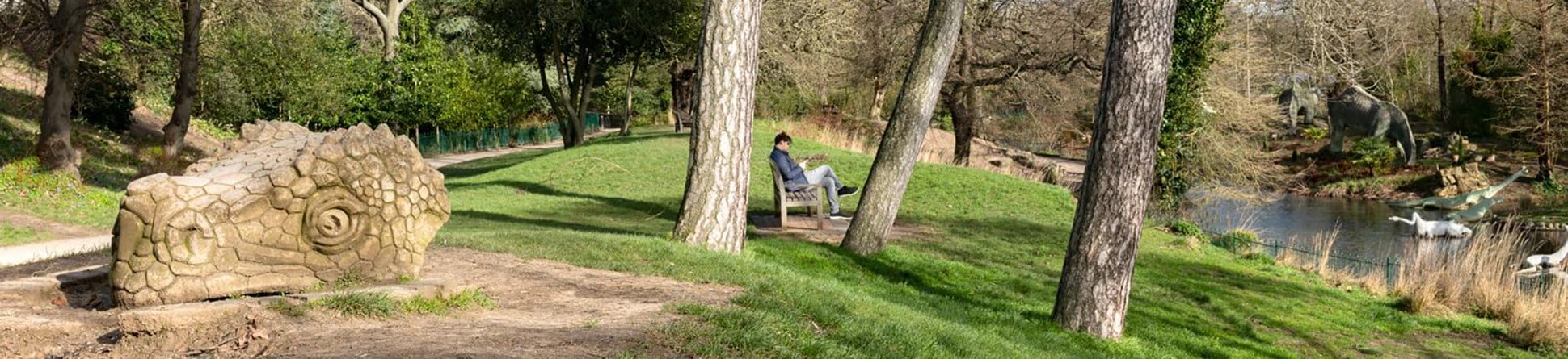 A man sits on a bench in parkland with dinosaur sculptures in front and behind him.