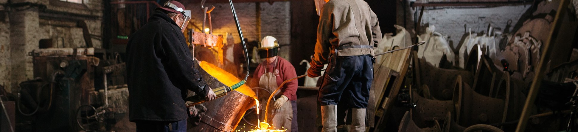 Three men pouring molten metal, demonstrating the bell casting process to a group of onlookers behind.