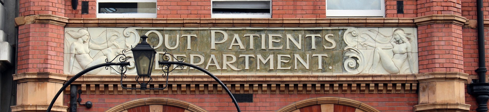 Entrance with the words 'Out Patients Department' engraved in the stone flanked by two women on either side. A lantern above the entrance gate partly obscures the view of the stone carving on the left