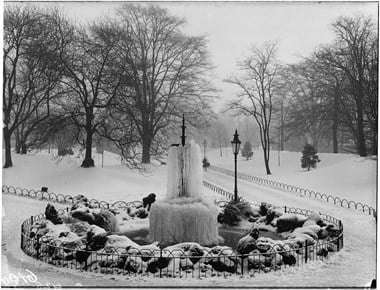 Snow covered parkland with bare skeletal trees. A fountain surrounded by snow covered shrubs/rocks is in the foreground.