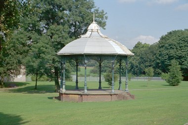 A bandstand in the foreground, surrounded by lawns with trees in the background.
