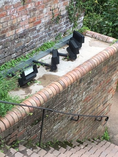 a set of four external lights on an elevated, flat concrete rectangular area. The area is encased with brick work with a brick wall in the background and brick stairs and an iron rail in the foreground