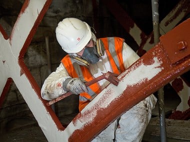 worker in white overalls, white safety hat and bright orange/grey jacket painting an iron structure in an interior setting