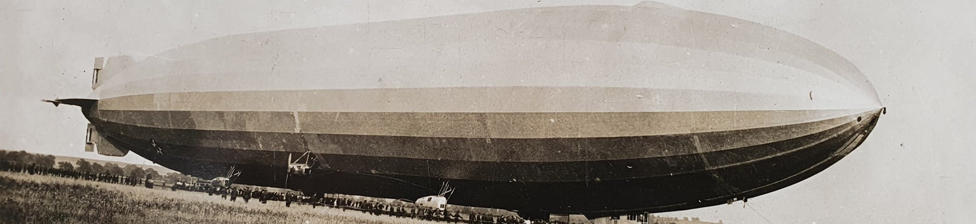 Black and white photo of Airship R.38/ZR 2 preparing for flight. The foreground is flat grassland. The airship sits on the horizon which has a line of dark specks along its entire length. Looking closer, the specks are people, some holding ropes. Hand written text on the bottom edge reads: "R.38. Ready for flight at Howden"