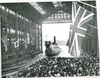 Crowds of people watching a submarine being launched down a slipway. British and Canadian flags are flying on either side