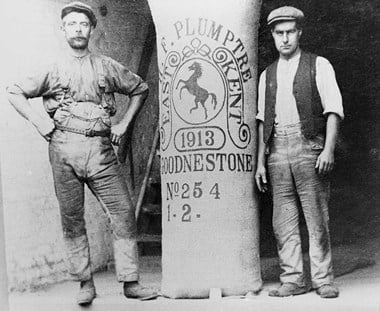 Two men in work clothes and flat caps standing either side of a pocket of hops which is taller than them