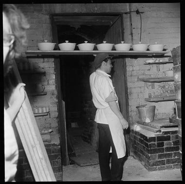 A man carrying a plank of dishes on his head at a pottery in the 1960s.