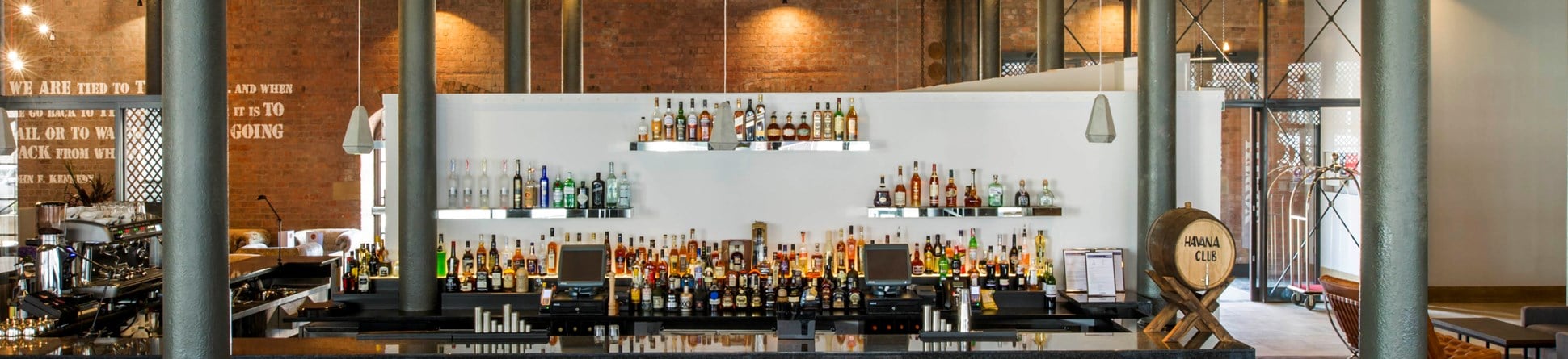 Chairs lined up at a bar in a high-ceilinged room with iron pillars and a red brick wall.