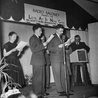 A group of people performing on stage under the banner which reads: 'RADIO BALONEY PRESENTS THE Lets At It Now Gang; Red light on when broadcasting