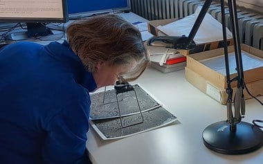 Photograph showing a short-haired woman in blue jumper leaning over a desk to look at an image through a magnifying device.