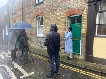 A film crew filming a woman dressed in period clothing outside her front door wearing a cleaning overall and smoking a cigarette.