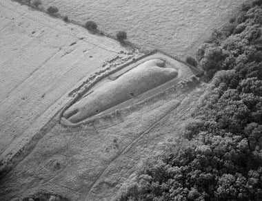Black and white oblique aerial photograph of long barrow set diagonally in the centre of the image. Trapezoid in shape, the barrow tapers towards the left. At the wider end is a wide, curved recess. At the opposite end is a narrow, elongated recess. The barrow is enclosed by a stone wall and is bordered by grassy fields, a field of cereal and dense woodland.