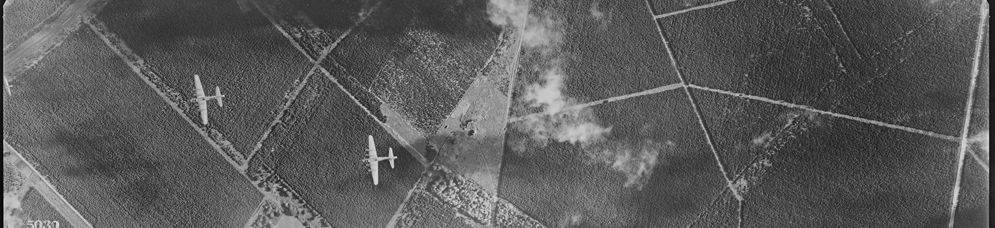 A black and white vertical aerial photograph of three military aircraft flying over a patchwork of plantations. Clouds obscure parts of the photograph and cast shadows on the ground.