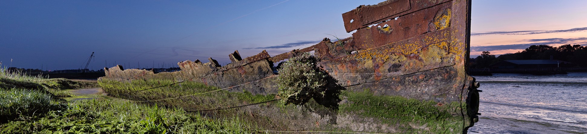 A rusting iron-shod ship hull stands on a shore, with vegetation in the foreground and a water channel beyond. 